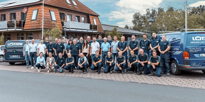 Eine Gruppe von Mitarbeitern steht vor einem Gebäude, umgeben von Fahrzeugen der Heizungs- und Sanitärfirma Lützow.