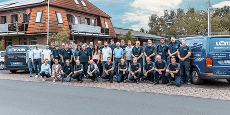 Eine Gruppe von Mitarbeitern steht vor einem Gebäude, umgeben von Fahrzeugen der Heizungs- und Sanitärfirma Lützow.