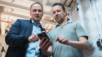 Zwei Männer stehen in einem Raum mit Rohren und schauen gemeinsam auf ein Smartphone, während sie etwas besprechen.
