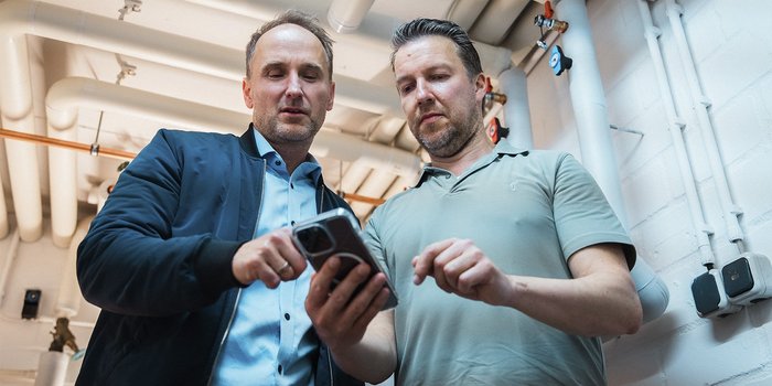 Zwei Männer stehen in einem Raum mit Rohren und schauen gemeinsam auf ein Smartphone, während sie etwas besprechen.