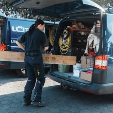 Ein Handwerker steht vor einem offenen Lieferwagen, während er ein langes Holzbrett in der Hand hält.