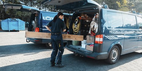 Ein Handwerker steht vor einem offenen Lieferwagen, während er ein langes Holzbrett in der Hand hält.