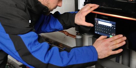 Ein Techniker in blauer Arbeitskleidung bedient ein digitales Bedienfeld einer Heizungsanlage mit beiden Händen.
