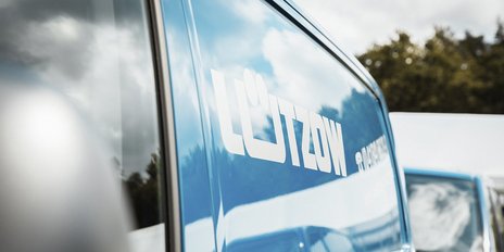 Ein blauer Lieferwagen mit dem auffälligen Schriftzug 'Lützkow' auf der Seite, umgeben von einer bewölkten Himmelsszenerie.