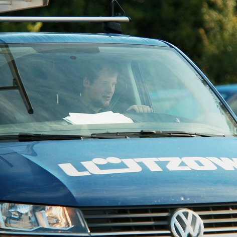 Ein Mann sitzt am Steuer eines blauen Lieferwagens mit einem Dachgepäckträger, während er auf die Straße schaut.