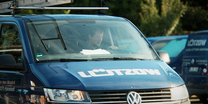 Ein Mann sitzt am Steuer eines blauen Lieferwagens mit einem Dachgepäckträger, während er auf die Straße schaut.