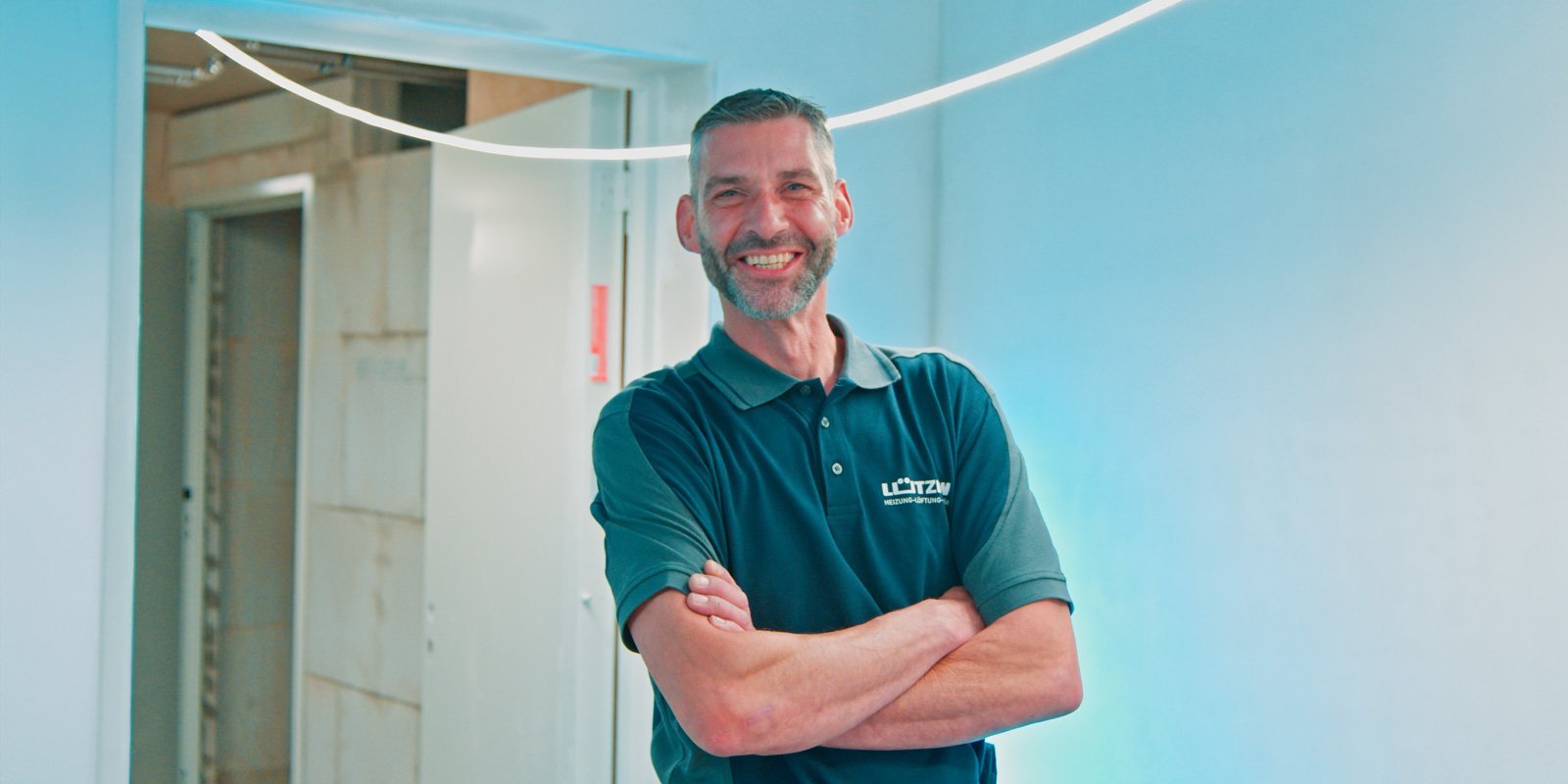 Ein lächelnder Mann mit Bart steht in einem hellen Raum und hat die Arme vor der Brust verschränkt.