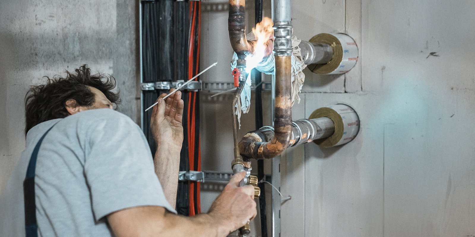 Ein Handwerker arbeitet an einer Rohrleitung, während eine Flamme aus einem Anschluss lodert, was auf eine Schweißarbeit hinweist.