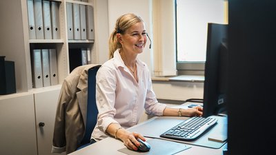 Eine lächelnde Frau mit Headset arbeitet konzentriert an einem Computer in einem modernen Büro mit Aktenregalen im Hintergrund.