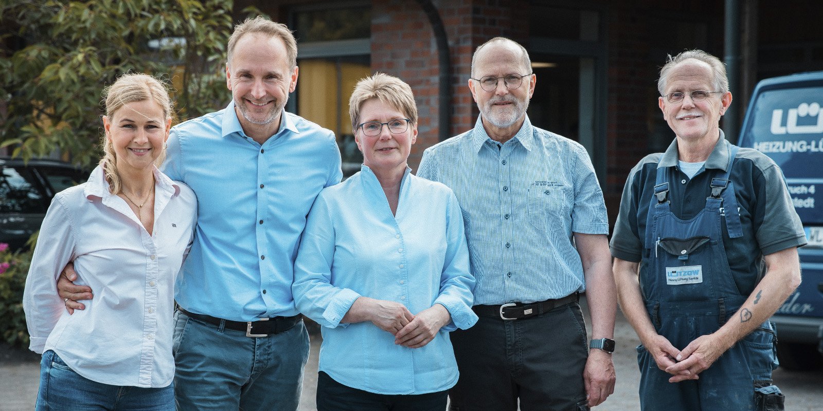 Fünf Personen stehen lächelnd vor einem Gebäude, alle tragen helle Hemden und zeigen eine freundliche Atmosphäre.