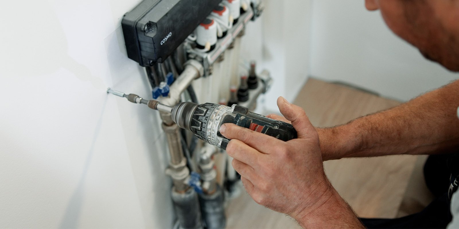 Ein Handwerker benutzt einen Akkuschrauber, um eine Schraube in eine weiße Wand zu drehen, während Rohre im Hintergrund sichtbar sind.