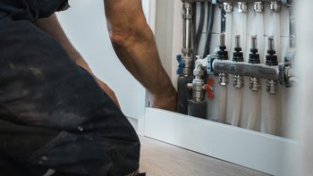 Ein Handwerker kniet und arbeitet an einer Heizungsanlage, während er mit Rohren und Ventilen in einem modernen Raum umgeht.