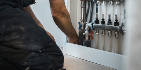 Ein Handwerker kniet und arbeitet an einer Heizungsanlage, während er mit Rohren und Ventilen in einem modernen Raum umgeht.