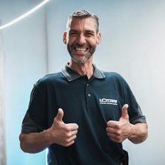 Ein lächelnder Mann in einem dunkelblauen Poloshirt zeigt Daumen hoch und strahlt Freude aus, während er in einem hellen Raum steht.