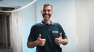 Ein lächelnder Mann in einem dunkelblauen Poloshirt zeigt Daumen hoch und strahlt Freude aus, während er in einem hellen Raum steht.