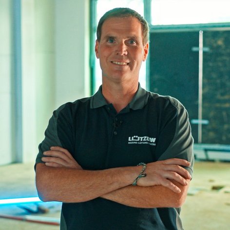 Ein Mann mit verschränkten Armen lächelt in einem hellen, unvollendeten Raum mit Baustellenmaterialien im Hintergrund.