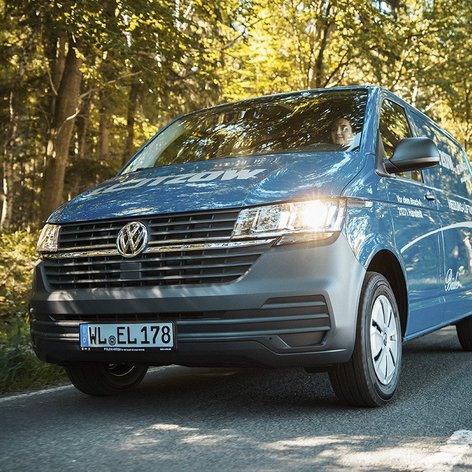 Ein blauer Volkswagen Transporter fährt auf einer ruhigen Landstraße, umgeben von üppigem, grünem Waldlicht.
