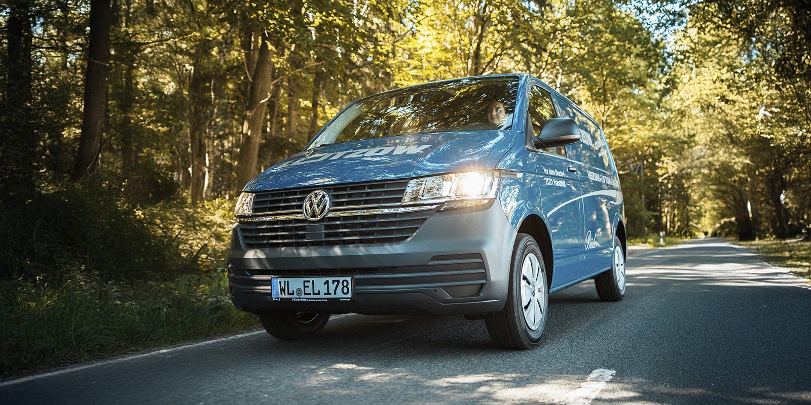 Ein blauer Volkswagen Transporter fährt auf einer ruhigen Landstraße, umgeben von üppigem, grünem Waldlicht.